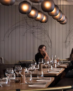 Smoke-grey Clérachic glass pendants in a long curving row above a restaurant, with a seated diner at a wooden table.