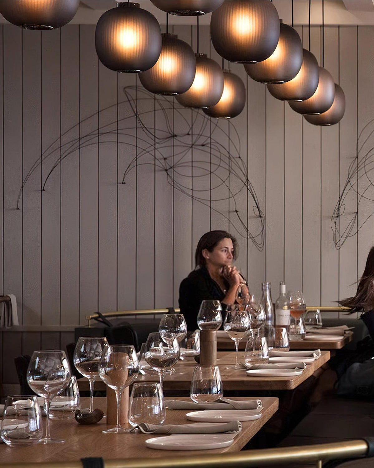 Smoke-grey Clérachic glass pendants in a long curving row above a restaurant, with a seated diner at a wooden table.