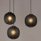Three smoke-grey Clérachic ribbed pendants glowing warmly, hung at staggered heights against a soft taupe backdrop.