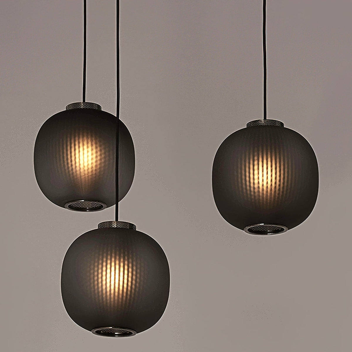 Three smoke-grey Clérachic ribbed pendants glowing warmly, hung at staggered heights against a soft taupe backdrop.