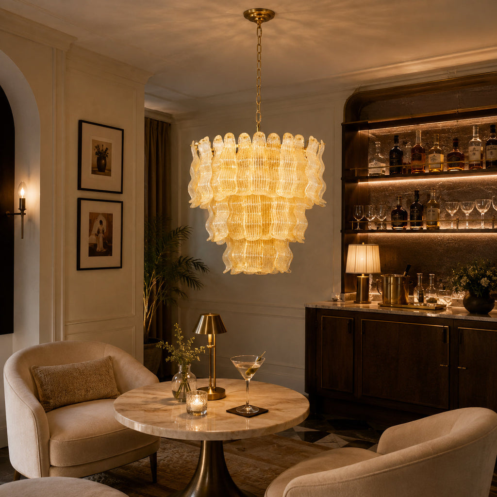 Lit yellow Clérachic pendant in a cosy lounge with a backlit bar shelf, a small marble table and two curved beige armchairs.