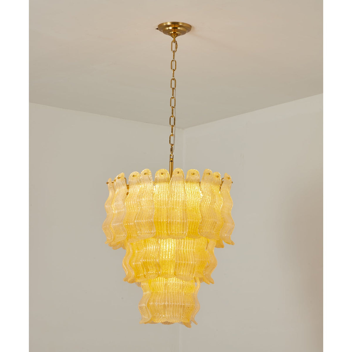 Lit yellow Clérachic pendant hung from a brass canopy and chain in the corner of a plain white room, its three tiers glowing.