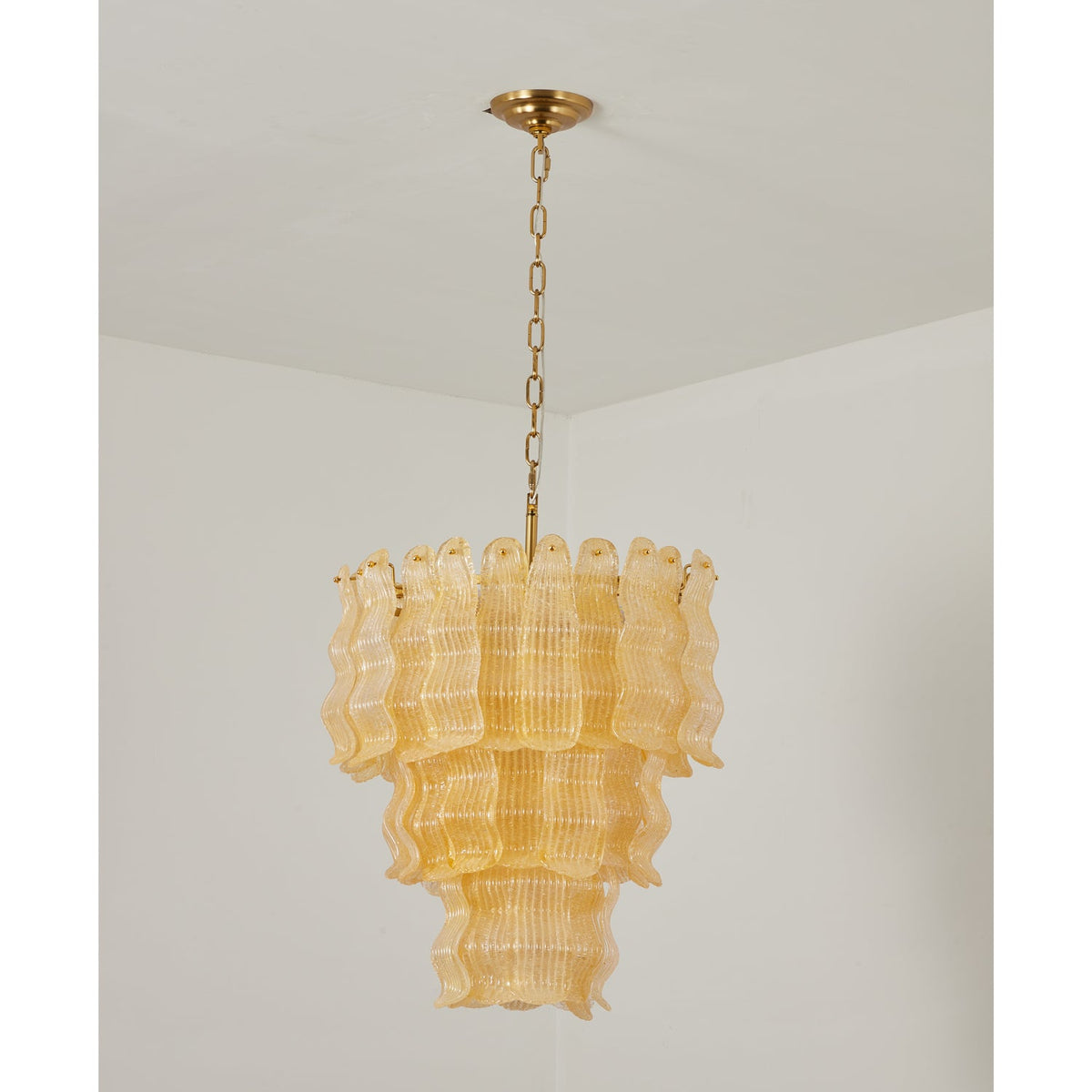 Unlit yellow Clérachic pendant with its pale amber ribbed glass tiers, hung from a brass chain against a white corner.