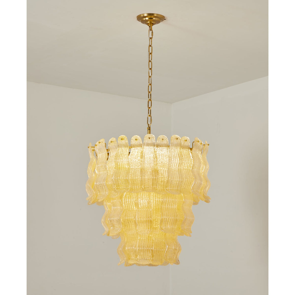 Lit Clérachic pendant in a softer yellow, three tiers of rippled glass panels on a brass chain in a white room.