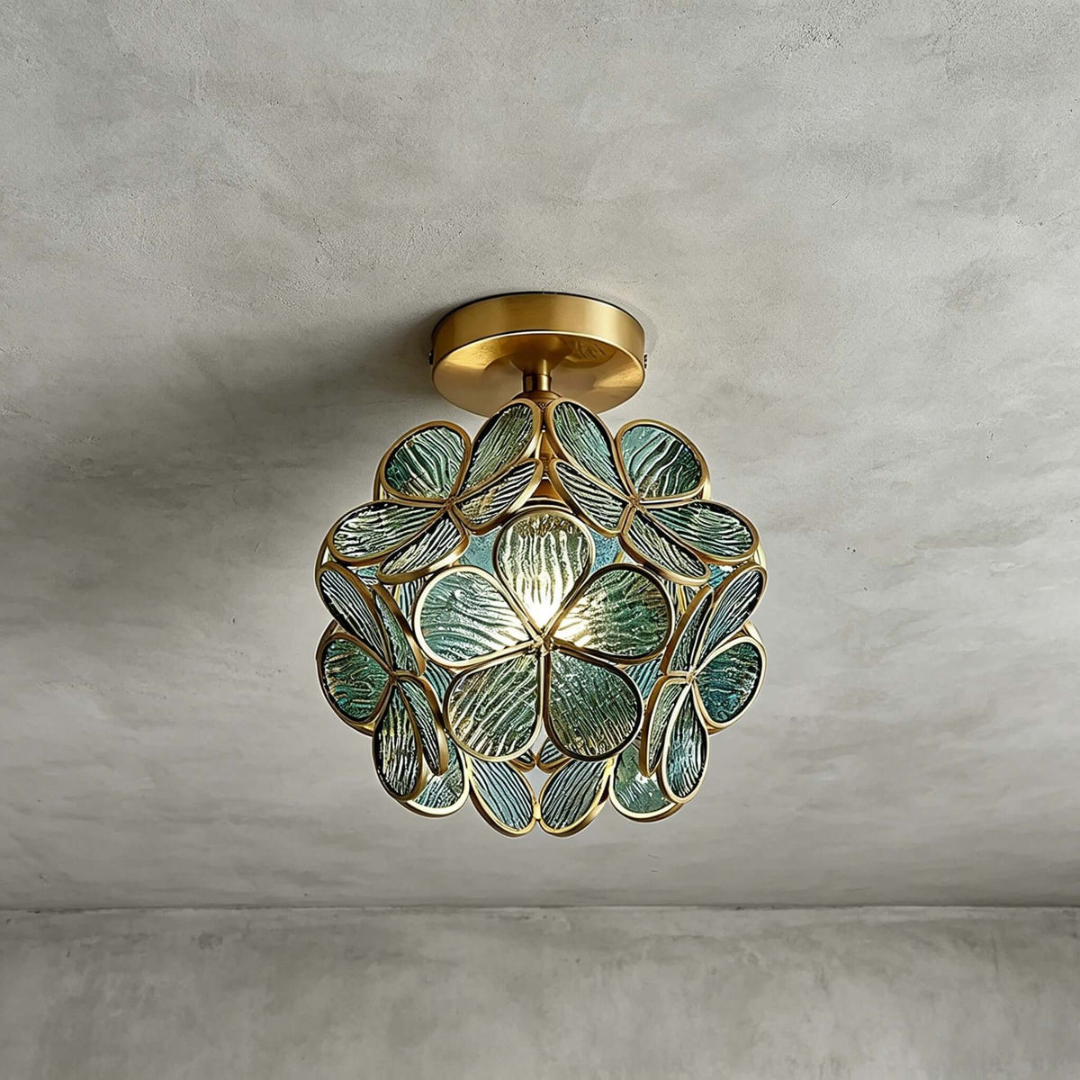 Blue-green glass-petal Clérachic ceiling light with a brass canopy, lit on a grey plaster ceiling.