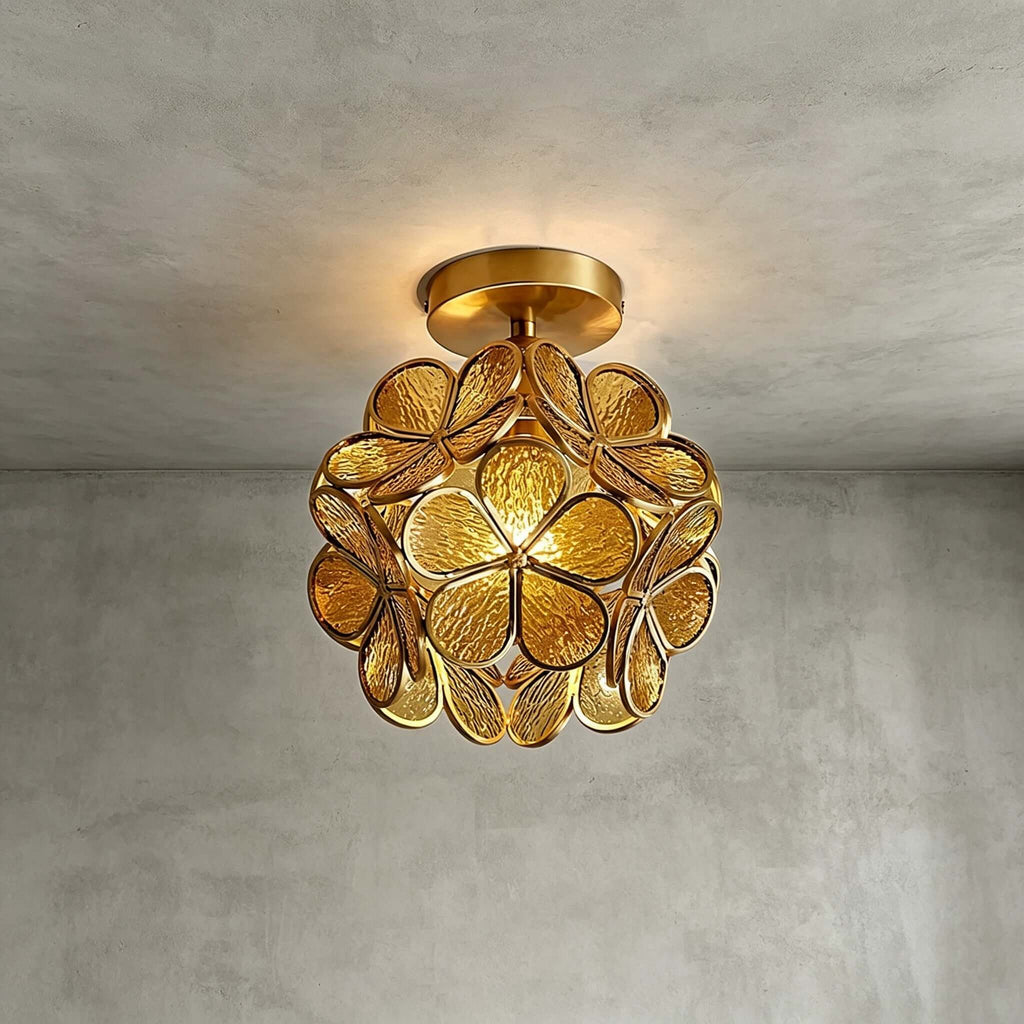 Honey-yellow glass-petal Clérachic ceiling light with textured petals and a brass frame, glowing on grey plaster.