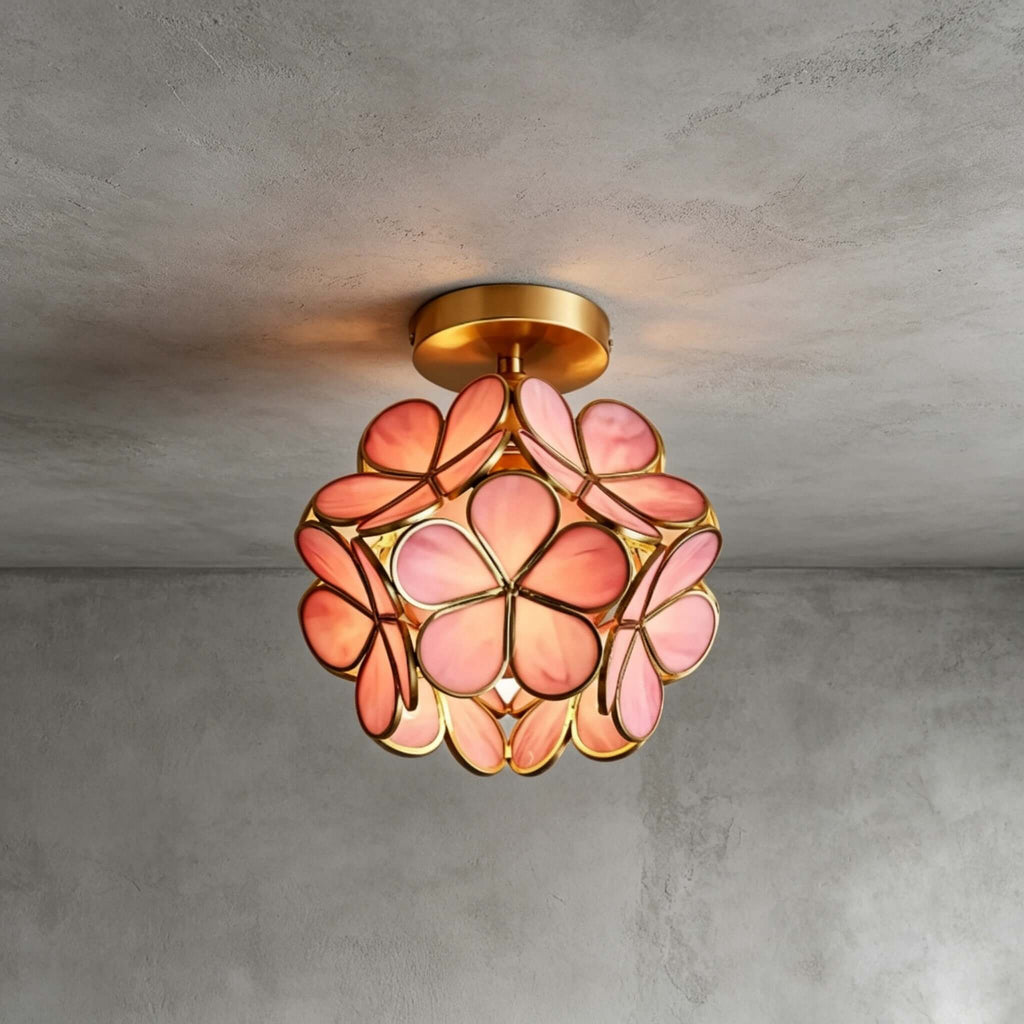 Pink glass-petal Clérachic ceiling light with a brass frame and canopy, lit against a grey plaster wall and ceiling.
