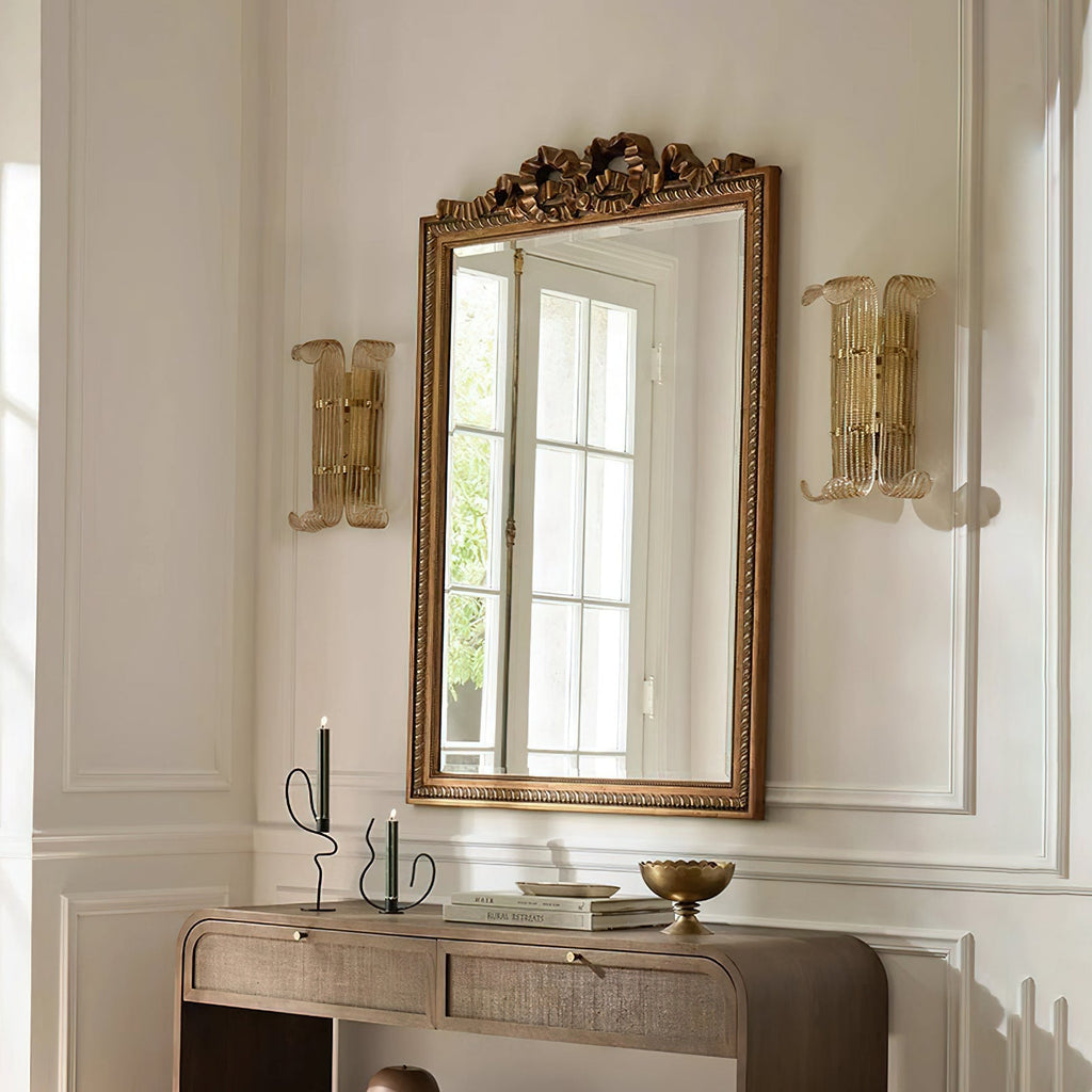 Two Clérachic wall lights in antique gold flank a gilt-framed mirror above a wooden console, with candlesticks on the console below.