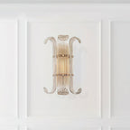 This Clérachic wall light hangs on a white panelled wall, its ribbed glass and curled arms glowing softly in warm light.