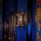 Unlit Clérachic glass-rod wall light mounted on a deep blue wall beside a black door, with colourful graffiti-style art at the edge.