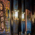 Two lit Clérachic wall lights glow warmly against dark blue walls, flanked by large colourful portrait paintings and a bar shelf below.