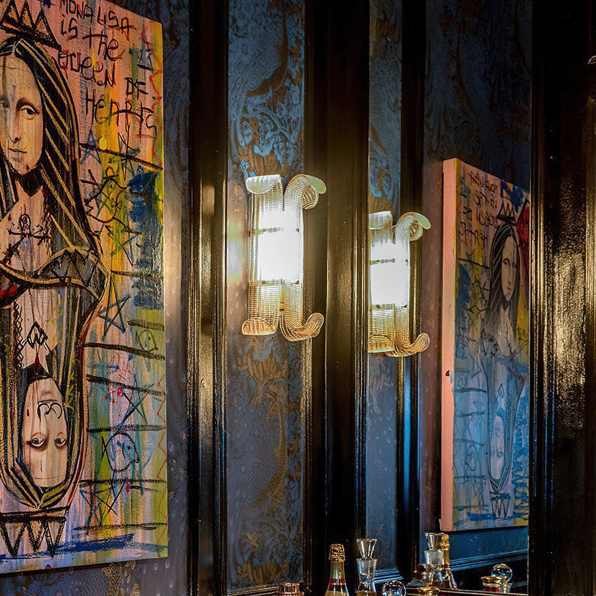 Two lit Clérachic wall lights glow warmly against dark blue walls, flanked by large colourful portrait paintings and a bar shelf below.