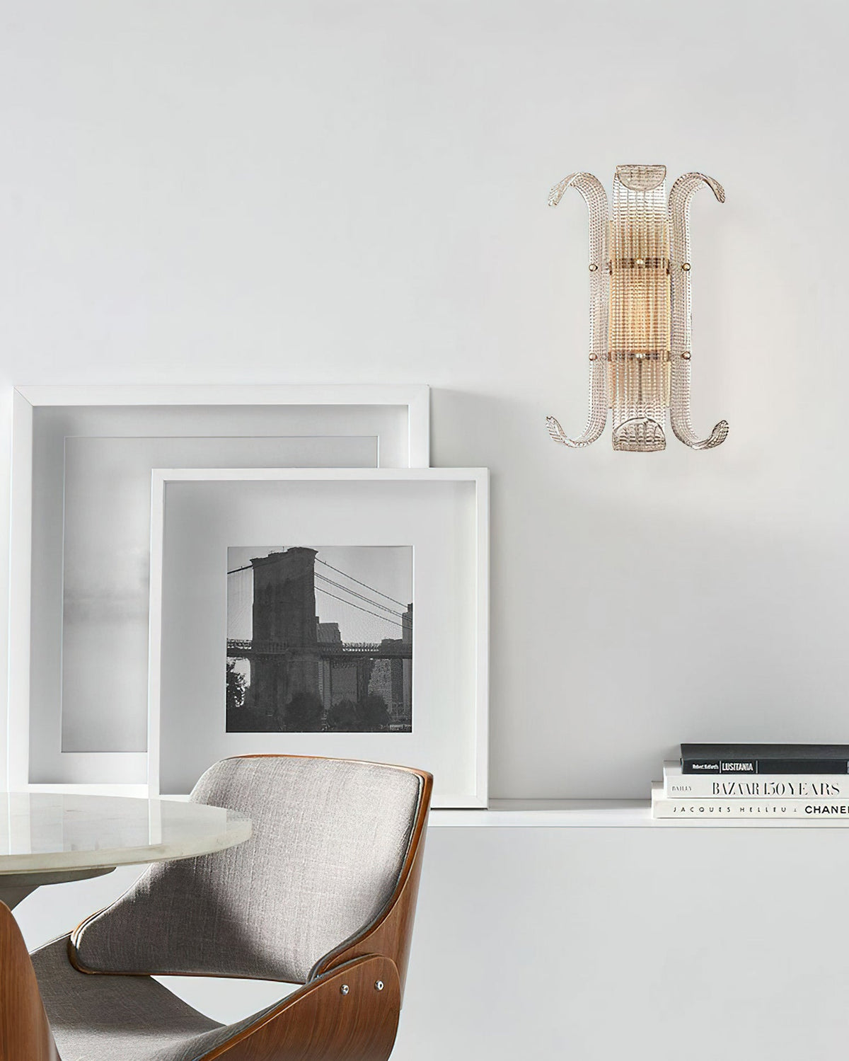 A Clérachic wall light on a pale grey wall above a corner with framed prints, a white shelf and an upholstered chair with a wooden frame.