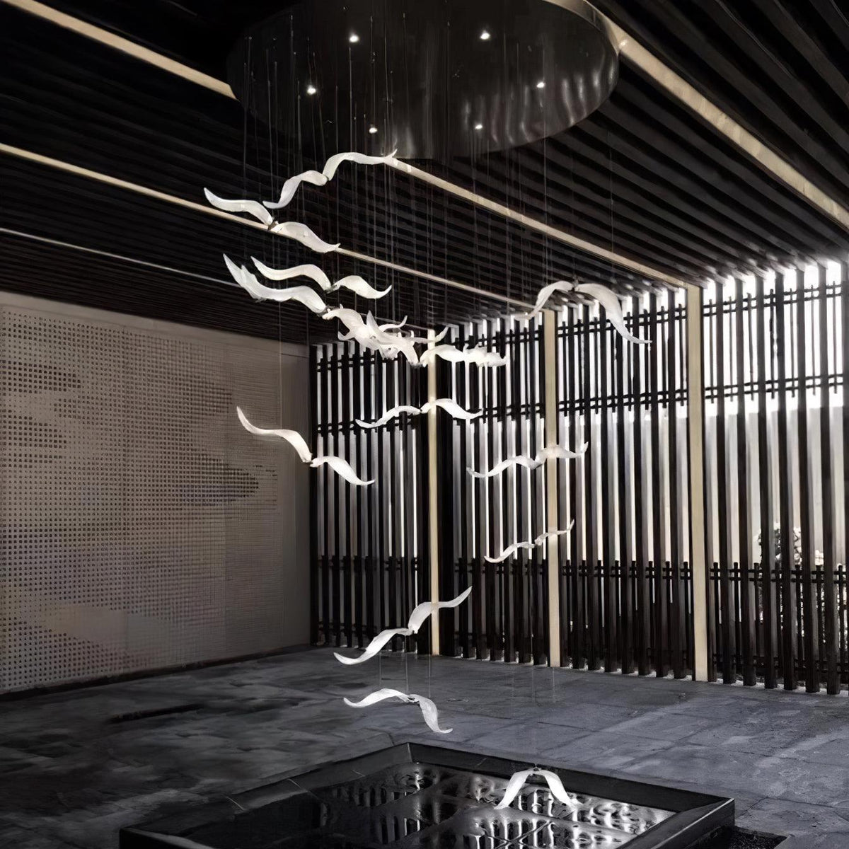 White glass gulls of this Clérachic chandelier hang on thin cables in a dark hall with slatted screens and a reflecting pool.