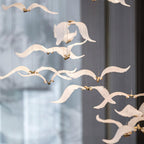Close view of white glass seagulls on this Clérachic chandelier, each with a small gold body, in soft focus against a grey window.