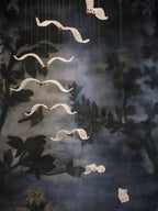 Glass gulls of this Clérachic chandelier hang in a vertical line in front of a dark painted wall of leaves.