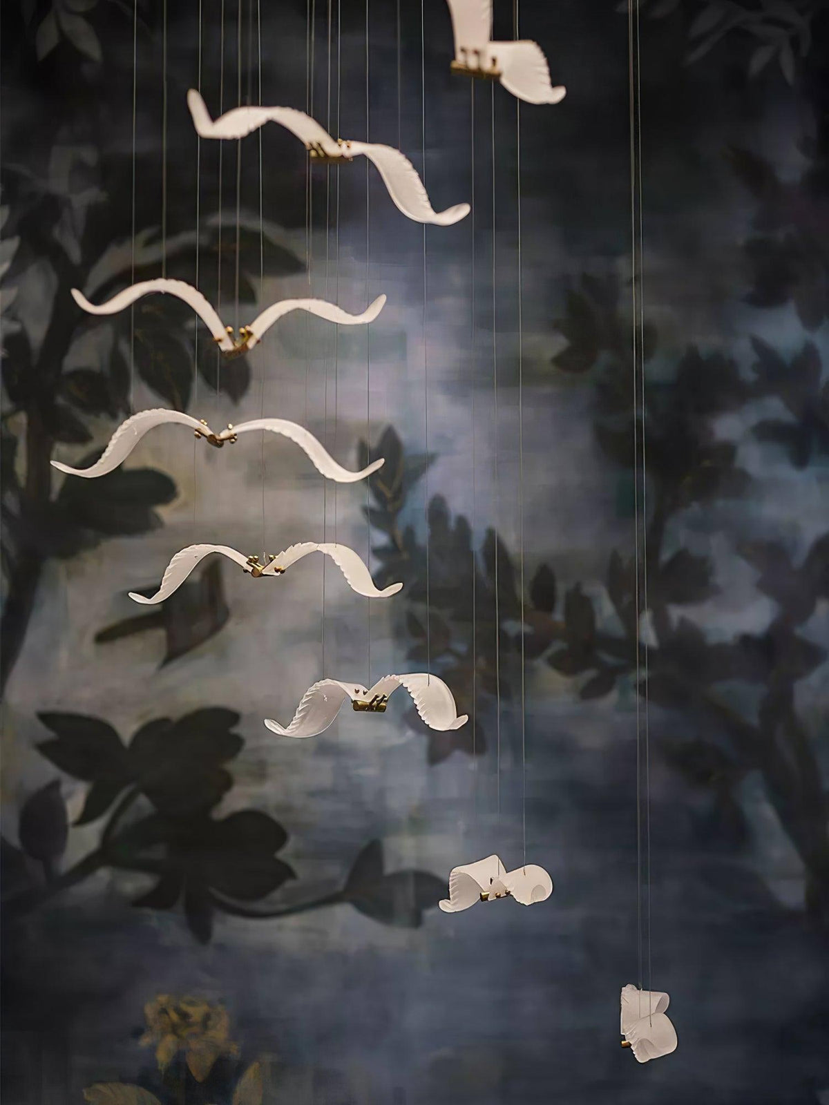 Glass gulls of this Clérachic chandelier hang in a vertical line in front of a dark painted wall of leaves.