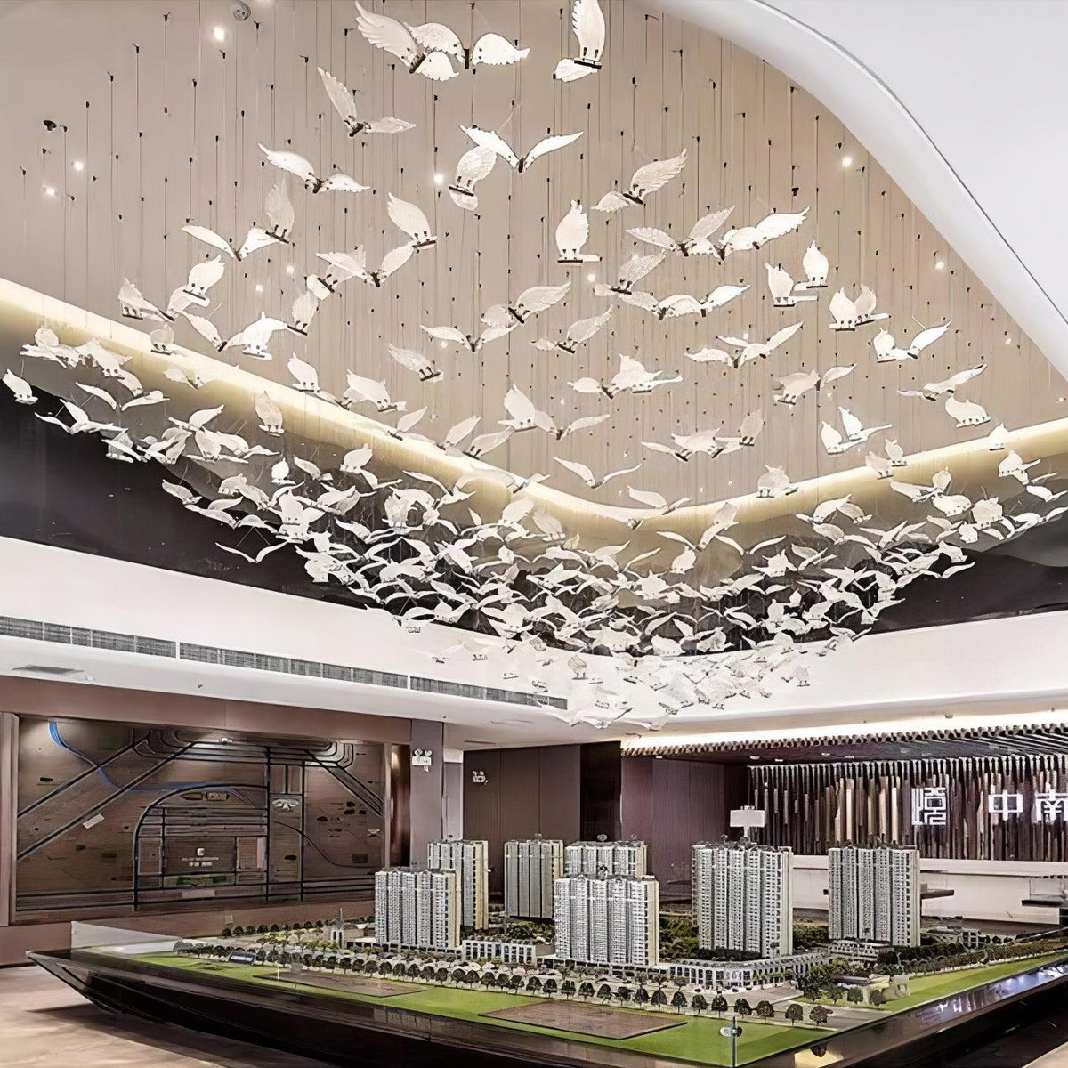 A large flock of Clérachic glass seagulls hangs under a curved ceiling in a showroom, above a landscape scale model.