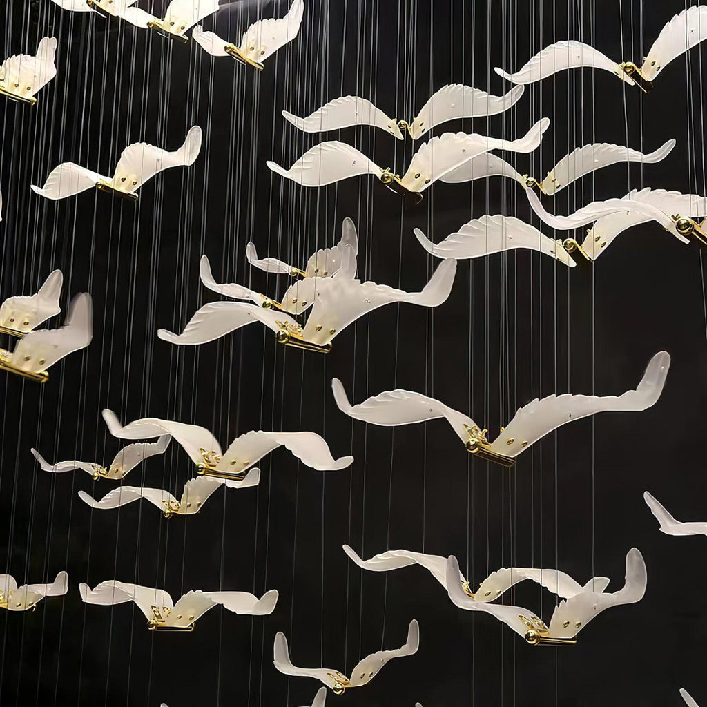 Close view of white glass seagulls with brass bodies on this Clérachic chandelier, hanging on fine cables against a black wall.