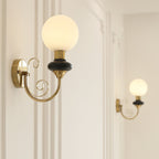 Close view of a Clérachic wall sconce with a black collar and glowing white globe, its gold arm ending in two scrolls.