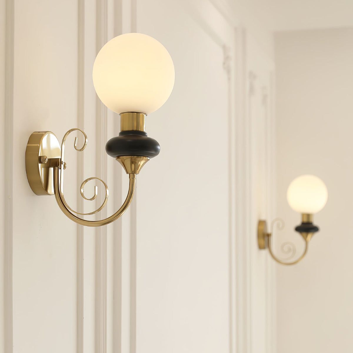 Close view of a Clérachic wall sconce with a black collar and glowing white globe, its gold arm ending in two scrolls.