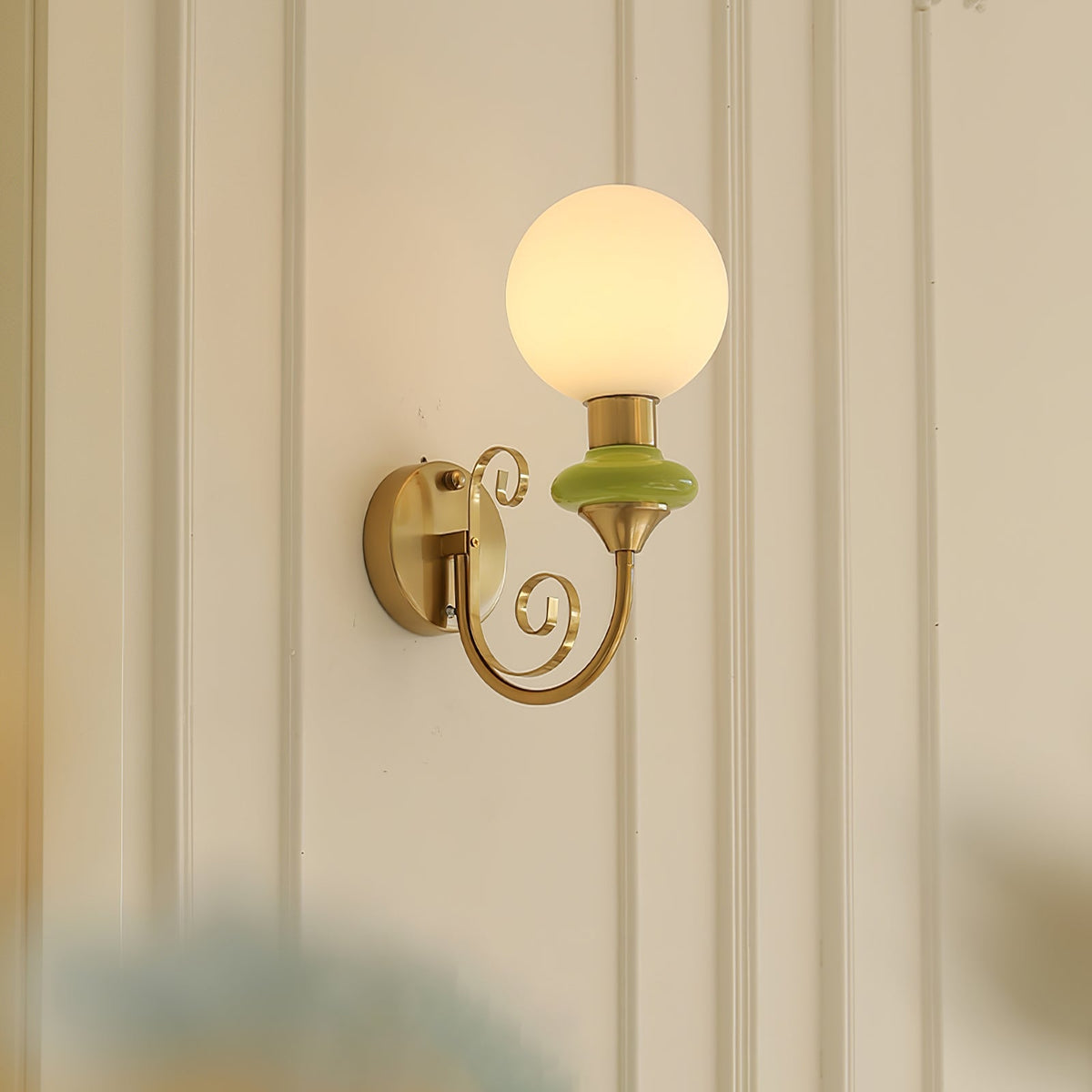 Green-collared Clérachic wall light glowing on a cream wall with vertical panelling, its gold backplate and scrolled arm in profile.