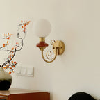 A Clérachic wall light with a mahogany collar on a cream wall above a wall switch, with autumn leaves and a wooden cabinet at left.