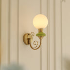 Green-collared Clérachic sconce on a white wall with moulded panels, the globe lit warm and the gold arm curling below.