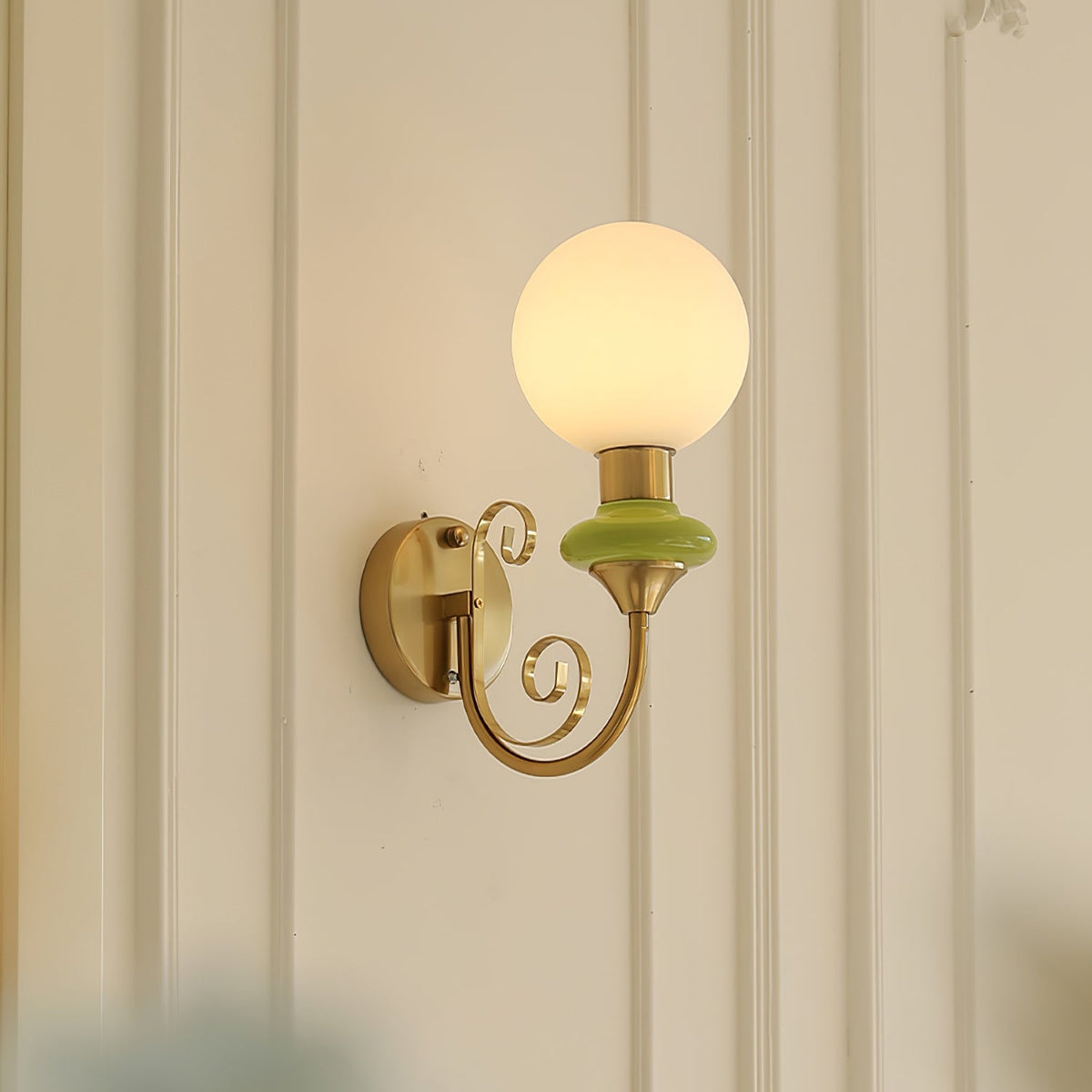 Green-collared Clérachic sconce on a white wall with moulded panels, the globe lit warm and the gold arm curling below.