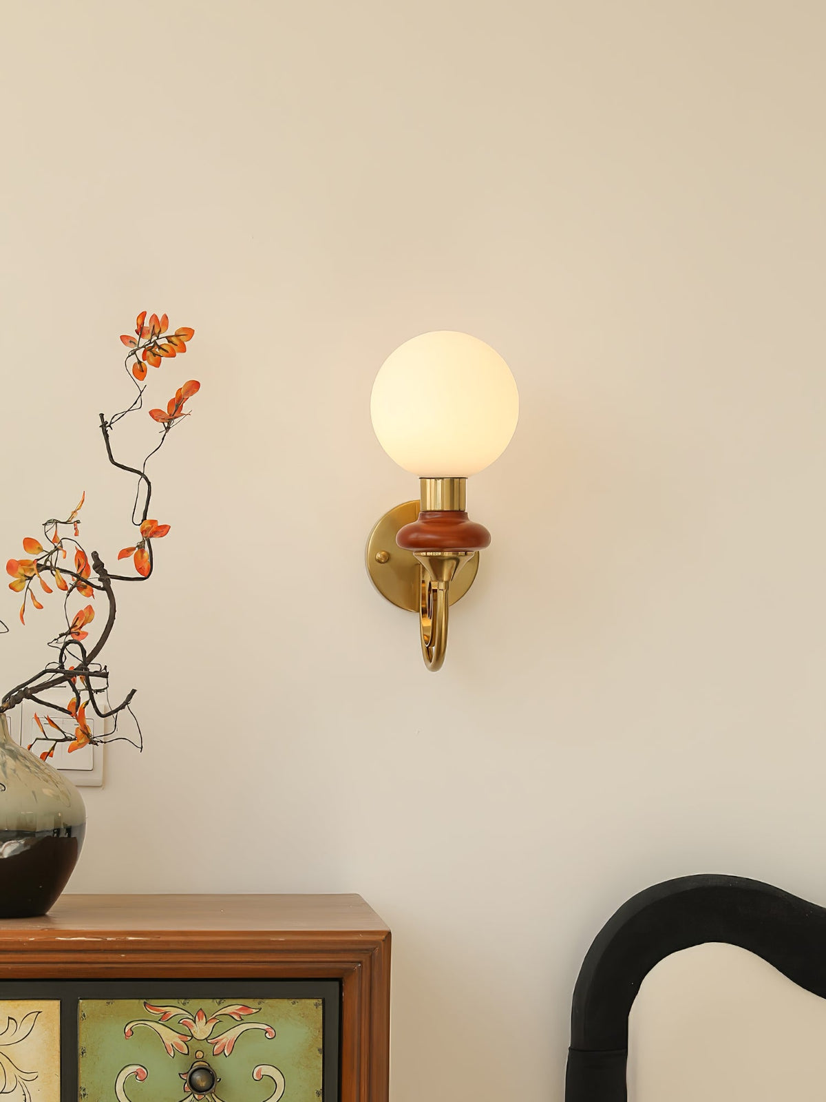 Clérachic wall light with a mahogany collar mounted above a painted wooden cabinet, with a branch of orange leaves beside it.