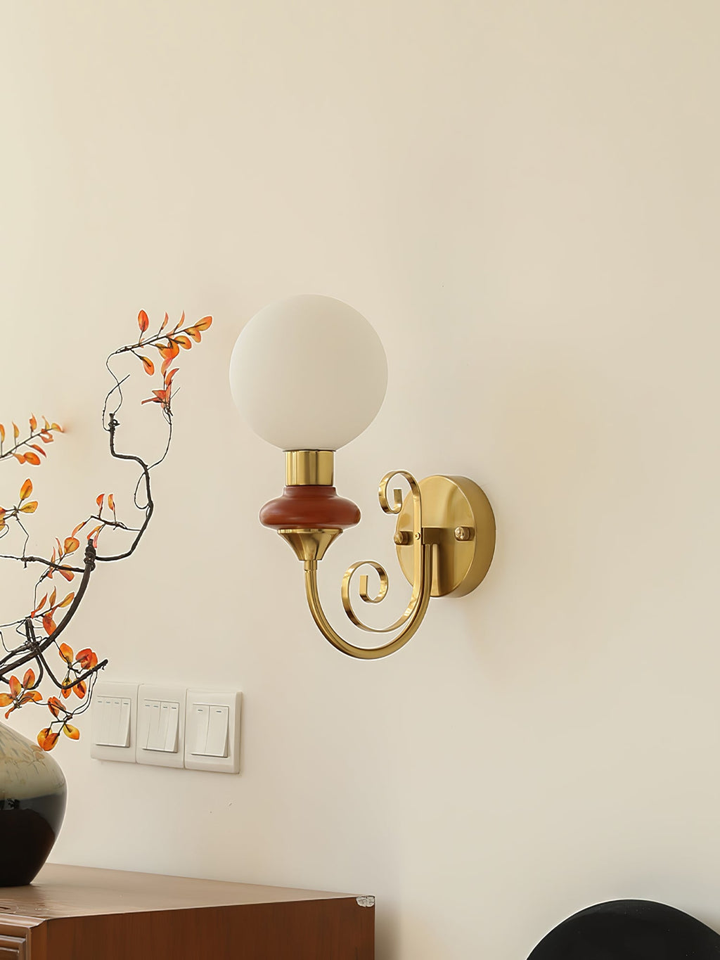 Clérachic wall light with a mahogany collar and unlit white globe on a cream wall, above a row of switches and a dark vase.
