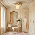 A Clérachic sphere ceiling light over a dressing table with a gilt mirror, a cushioned stool and fitted wardrobes in cream.