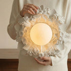 Someone in a cream knit jumper holds this Clérachic ceiling light, showing its petalled resin collar and lit glass sphere up close.