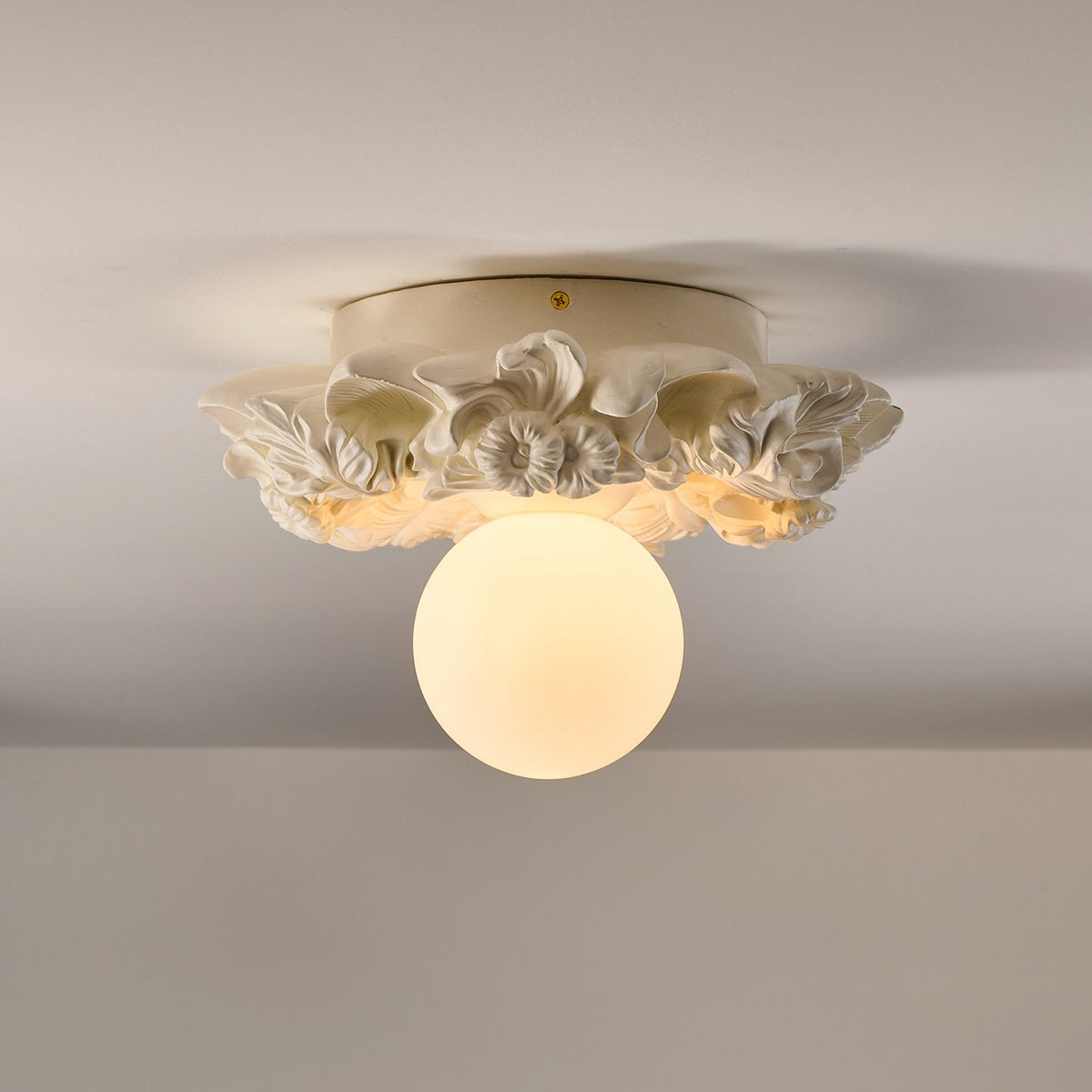 Ivory Clérachic ceiling light on a grey ceiling, its carved flower collar and frosted sphere glowing softly.