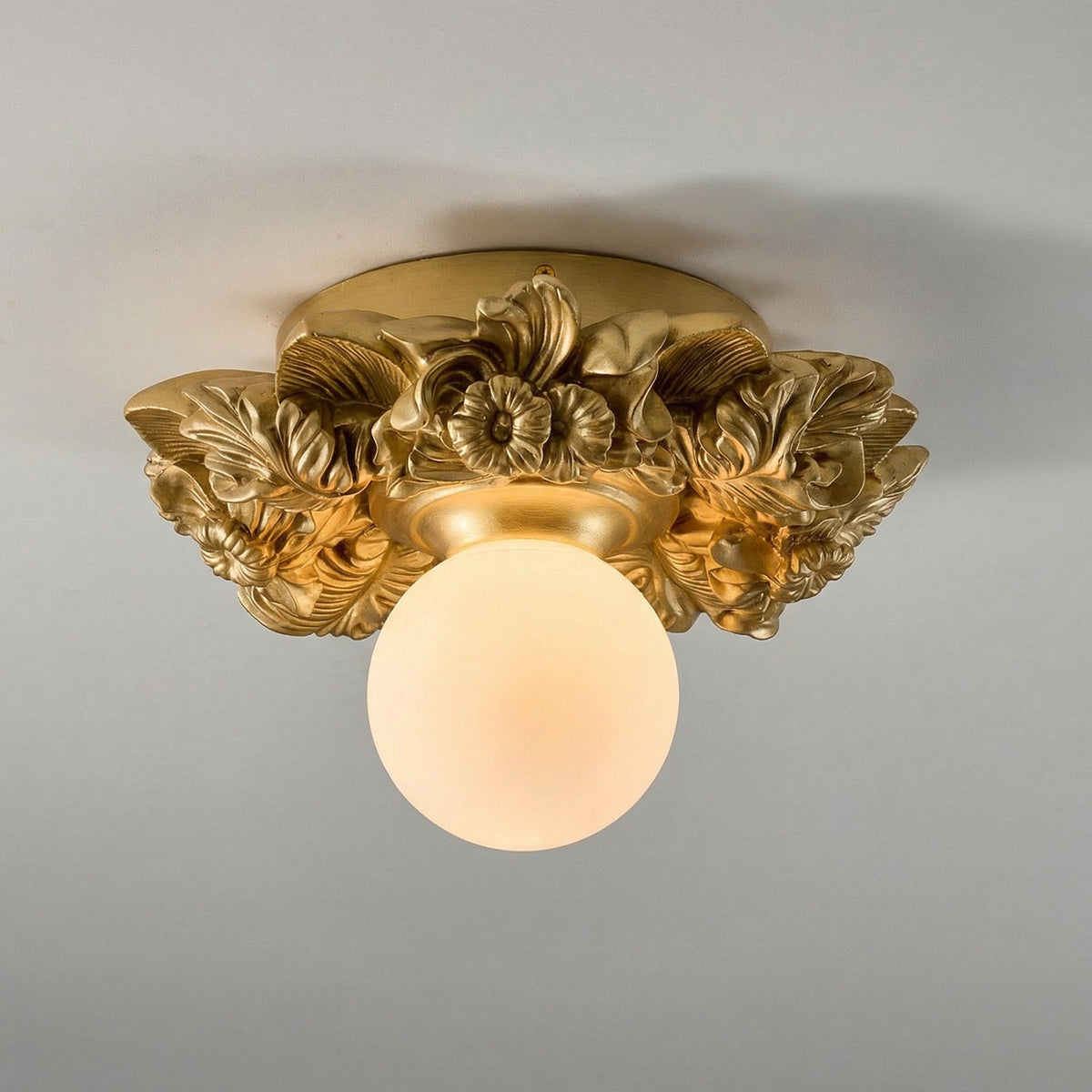 Gilded gold version of this Clérachic ceiling light on a grey ceiling, with a warm-lit frosted glass sphere below the flower collar.