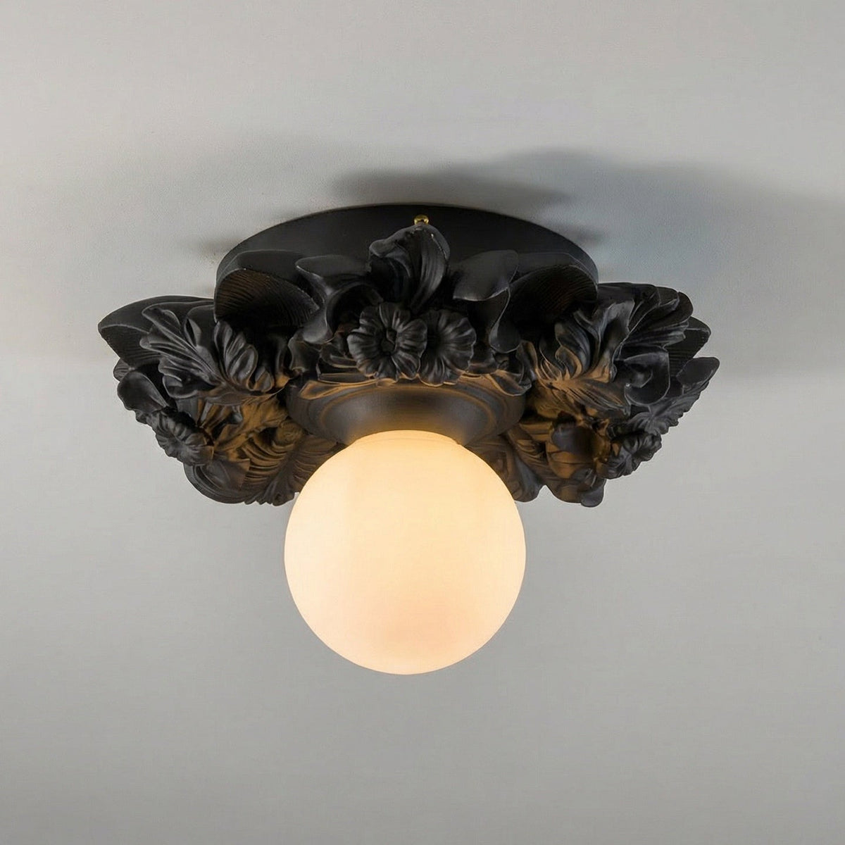 Matte black version of this Clérachic ceiling light on a grey ceiling, its dark resin flowers framing a warm-lit glass sphere.