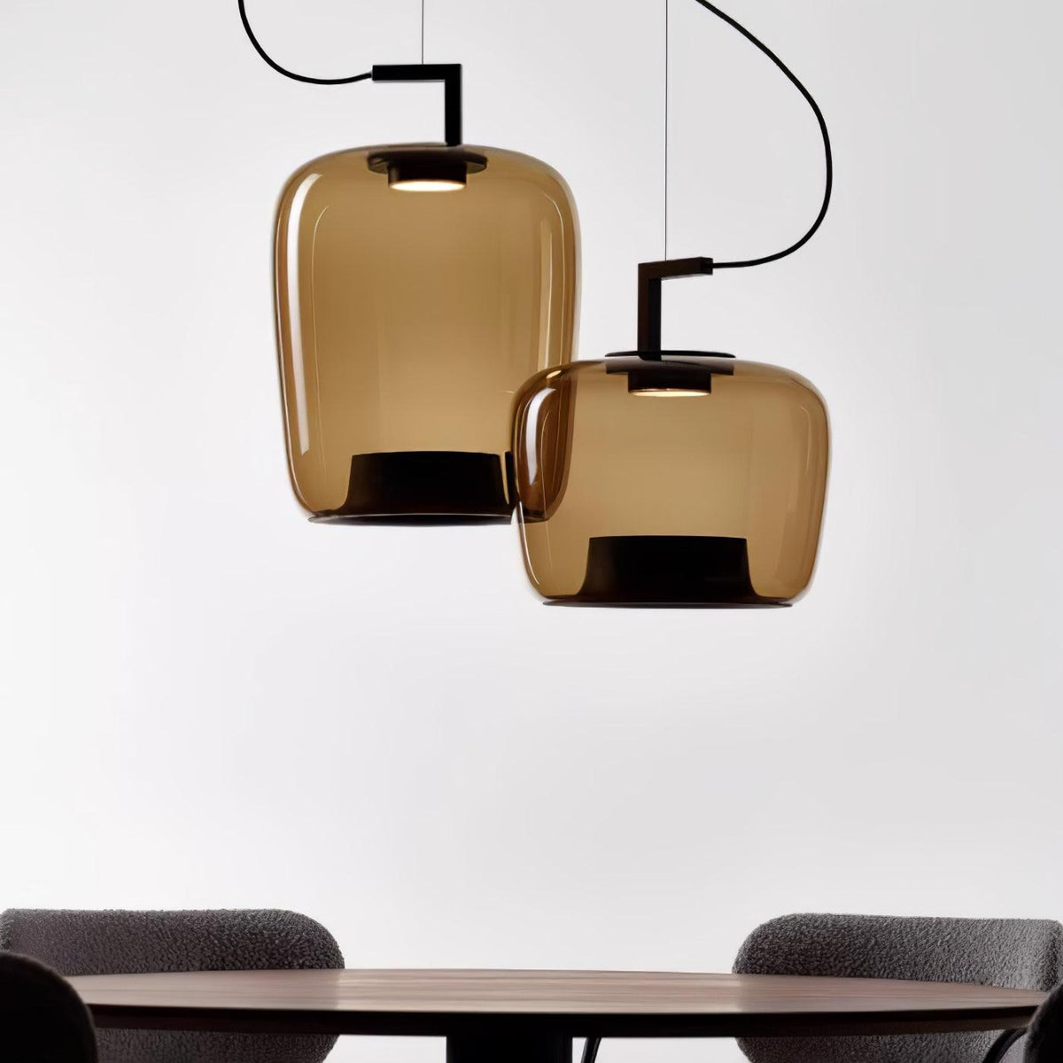 Close crop of two amber glass Clérachic pendants, one tall and one squat, above a walnut table edge and grey chairs.
