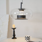 Clear glass Clérachic pendant with a black inner collar, glowing above a black candlestick and a checked mug on a white table.