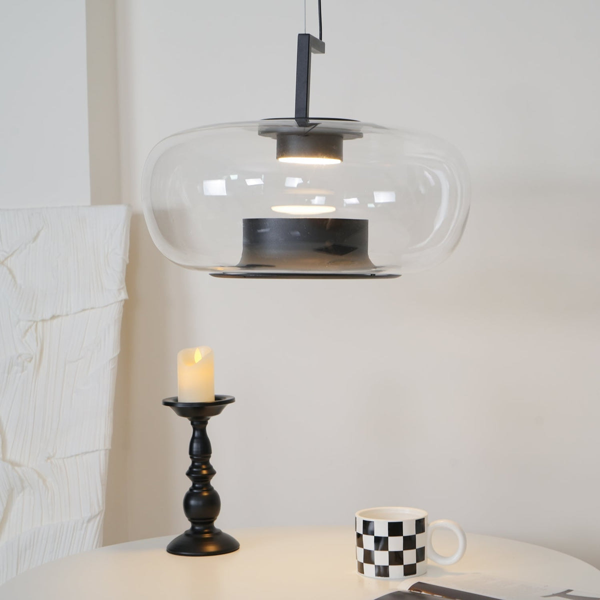 Clear glass Clérachic pendant with a black inner collar, glowing above a black candlestick and a checked mug on a white table.