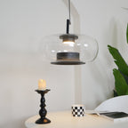 Wide clear glass Clérachic pendant seen from a slight angle above a candlestick, a checked mug and an open book.