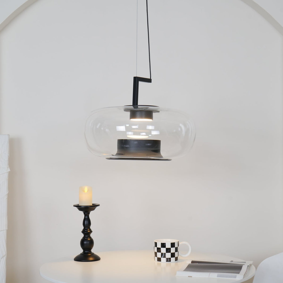 Clear glass Clérachic pendant with a black stem bracket over a candlestick, a checked mug and a magazine on a white table.