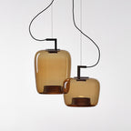 Two amber glass Clérachic pendants, one tall and one rounded, on separate black brackets against a pale backdrop.