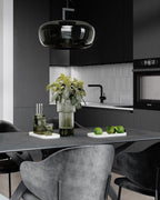 Wide smoke grey glass Clérachic pendant above a dark table set with a vase, glasses and limes, in a black kitchen.