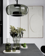 Close view of a smoke grey glass Clérachic pendant above a table with a ribbed vase, a carafe and limes.