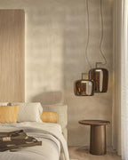 Two metallic amber Clérachic pendants hang beside a bed with a round wooden side table, against a patterned wall.