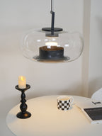 Lit wide clear glass Clérachic pendant above a candle, a checked mug and a magazine on a softly lit white table.