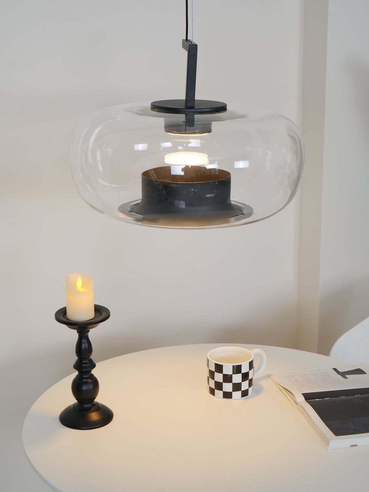 Lit wide clear glass Clérachic pendant above a candle, a checked mug and a magazine on a softly lit white table.