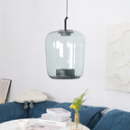 Tall grey-green glass Clérachic pendant in front of a white textured panel, above a table of books and a gold vase.