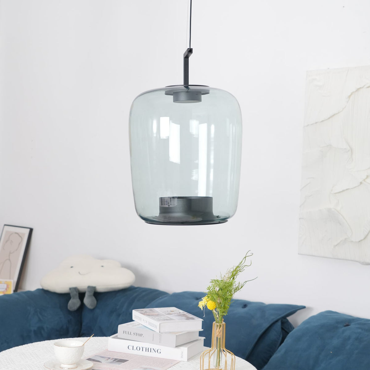 Tall grey-green glass Clérachic pendant in front of a white textured panel, above a table of books and a gold vase.