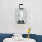 Tall grey-green glass Clérachic pendant against a textured white panel, above a table with books, a teacup and a yellow flower.